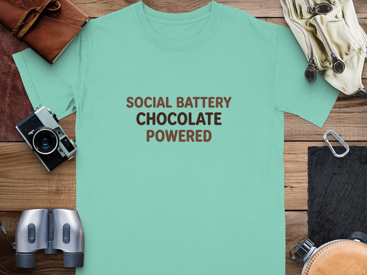 Teal t-shirt with "Social Battery Chocolate Powered" text, surrounded by travel items.